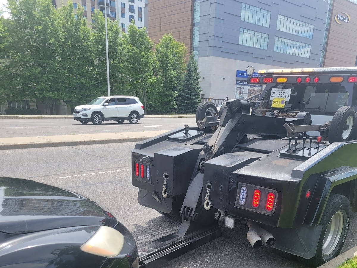 Rear view of a black heavy-duty tow truck towing a black car on a city street, with buildings, trees and a white SUV in the background.