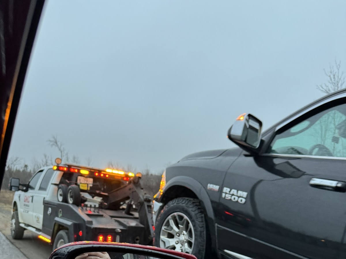 View from inside a vehicle of a tow truck with flashing emergency lights stopped beside a black Ram 1500 pickup truck on an overcast day